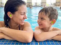 Mutter und Sohn stützen sich am Beckenrand eines Schwimmbads ab und lächeln sich an, beide tragen grüne Snugos Ohrstöpsel als Spritzwasserschutz.