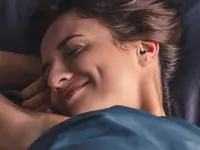 SweetDreams Nova – bequemer Schlaf-Ohrstöpsel für Seitenschläfer Lächelnde Frau liegt im Bett und trägt einen dunkelblauen Ohrstöpsel.
