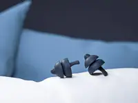 SweetDreams Nova – Schlaf-Ohrstöpsel mit SoftGrip auf Kissen Zwei dunkelgraue Ohrstöpsel mit Doppel-Lamellen auf weißem Kissen vor blauen Kissen.